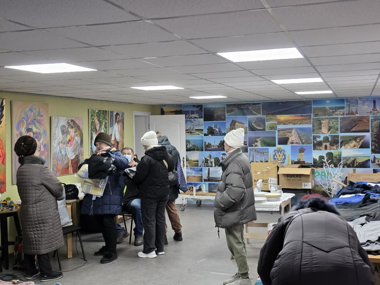 Група людей у теплій одежі зібралася у світлому приміщенні, стіни якого прикрашені художніми картинами та колажем із фотографій краєвидів; на передньому плані видно коробки та речі.