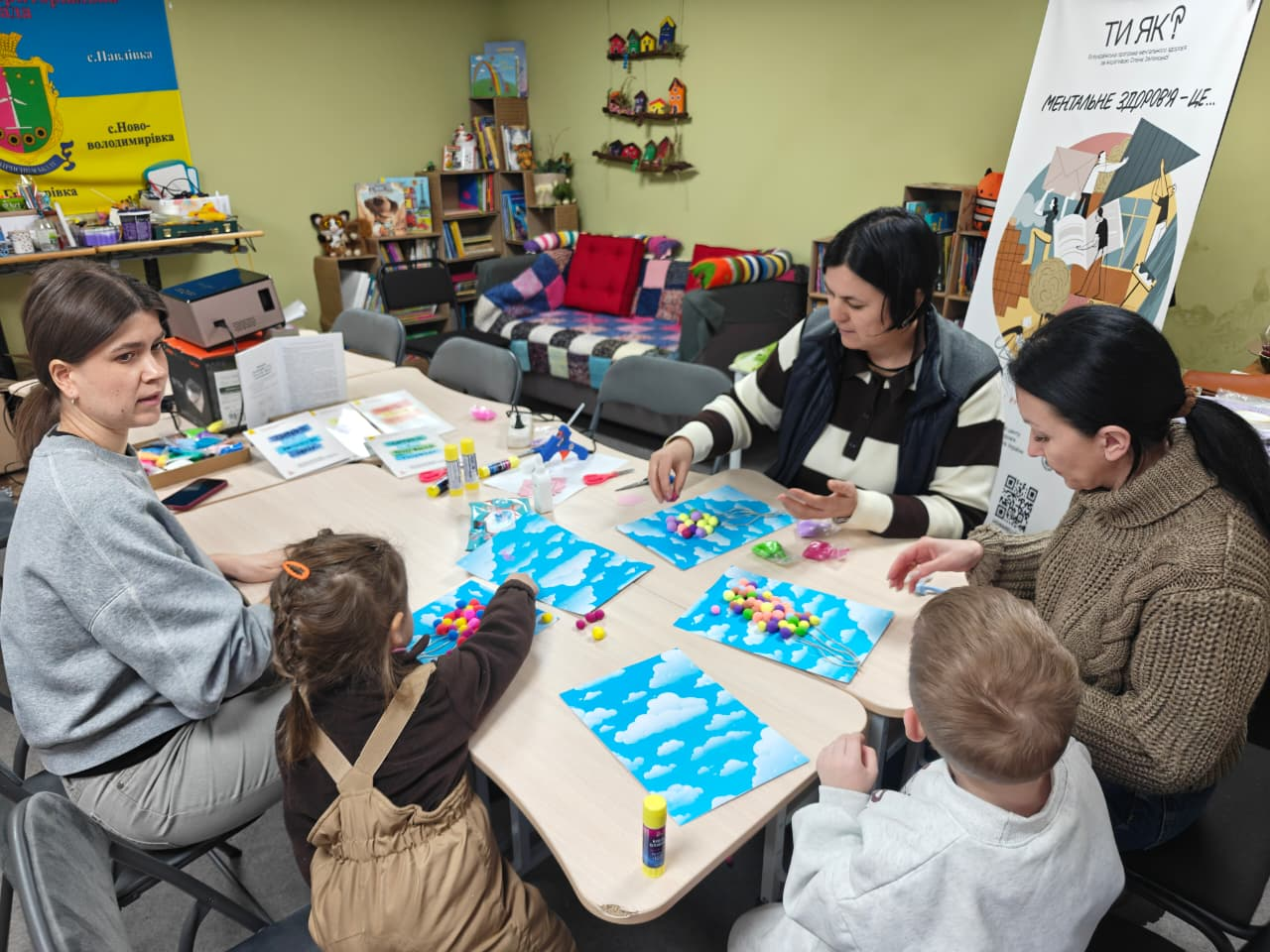 Дорослі та діти сидять за столом у затишній кімнаті, займаючись творчістю: вони створюють аплікації з кольорових помпонів на аркушах із зображенням неба; поруч стоїть інформаційний стенд про ментальне здоров'я.