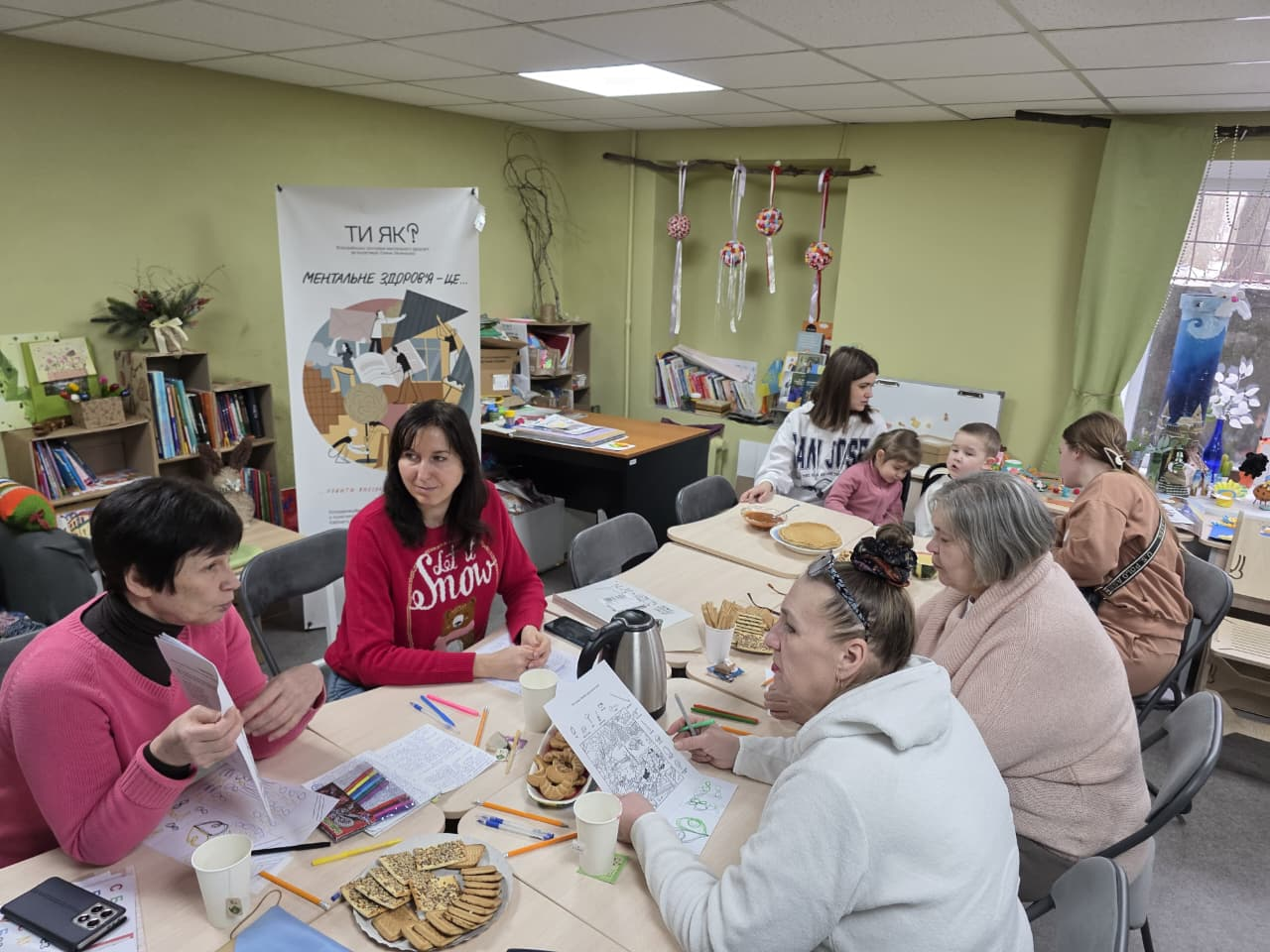 Група жінок та дітей зібралася за спільним столом, де вони спілкуються, розфарбовують малюнки та частуються печивом; на фоні видно книжкові полиці та плакат про ментальне здоров'я «Ти як?».