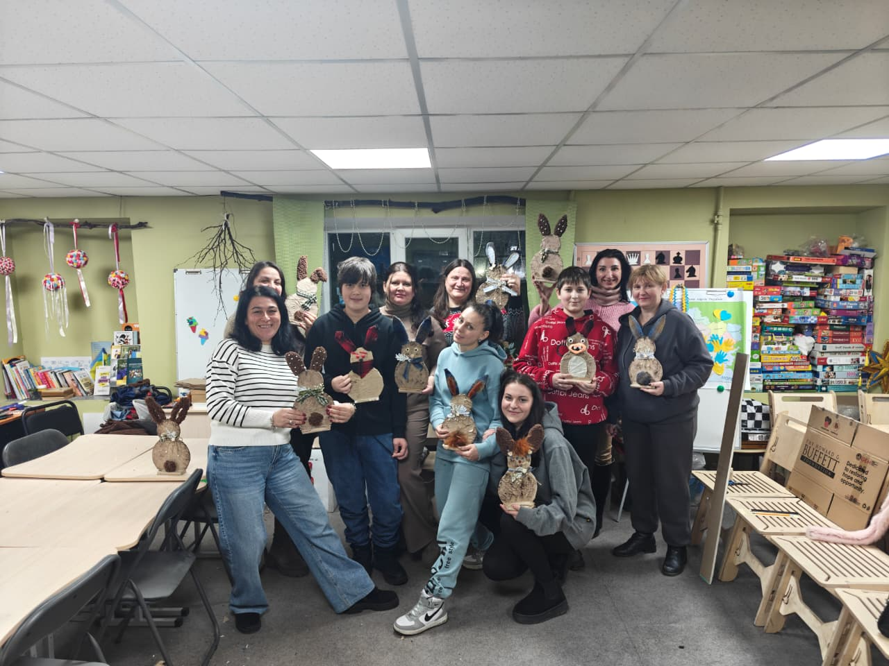 Група жінок та підлітків позує для спільного фото в ігровій кімнаті, тримаючи в руках саморобні вироби у формі зайчиків; на фоні видно полиці з іграшками та настільними іграми.