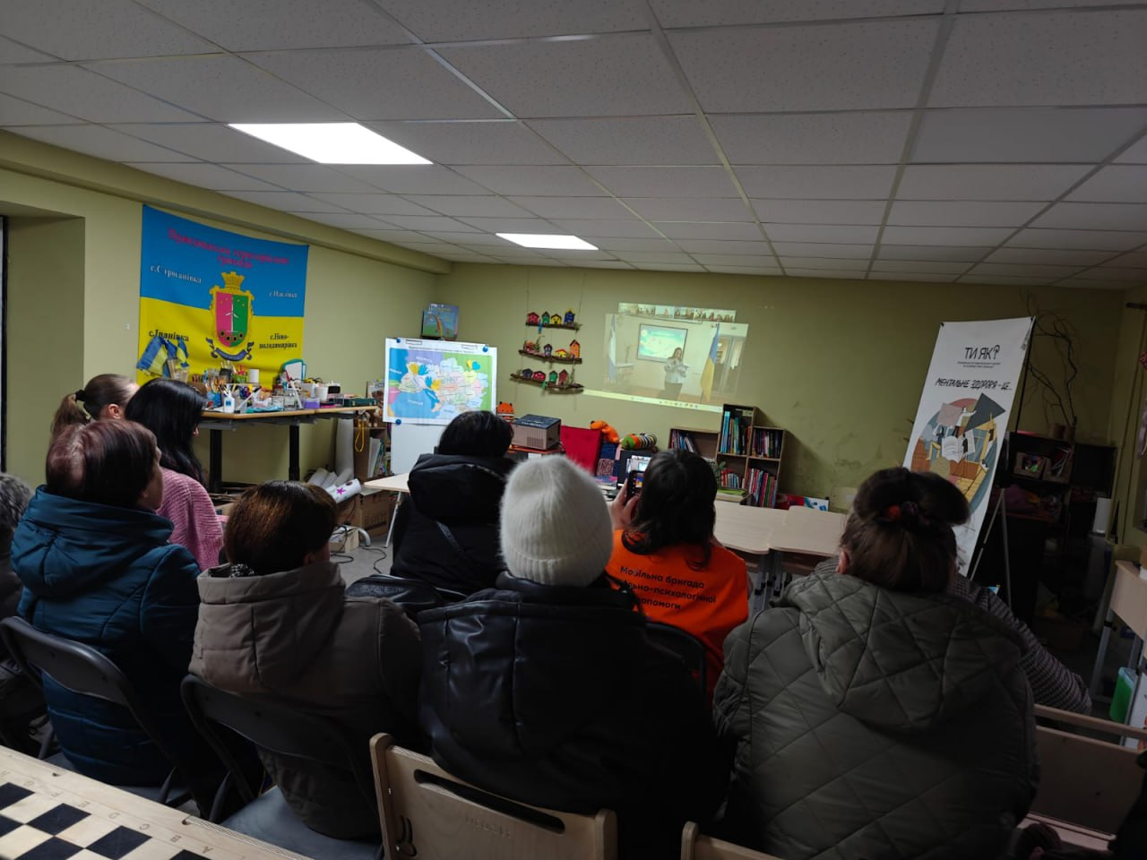 Люди в залі переглядають відеопрезентацію на стіні; у приміщенні розміщено карту України, національну символіку та стенд програми ментального здоров'я «Ти як?».