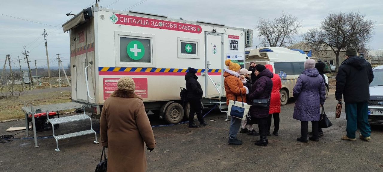 Мобільний медичний пункт з написом «Світлиця здоров'я CASERS» у сільській місцевості. Біля білого фургона на колесах та поруч розташованої машини швидкої допомоги стоїть група людей у зимовому одязі, які очікують на прийом.
