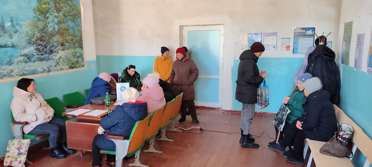 Група людей у зимовому одязі перебуває в приміщенні, схожому на приймальню або зал очікування. Кілька осіб сидять за столом та на стільцях уздовж стін, інші стоять. На стінах розміщені інформаційні плакати та пейзажна картина. Панує спокійна атмосфера очікування або консультації.