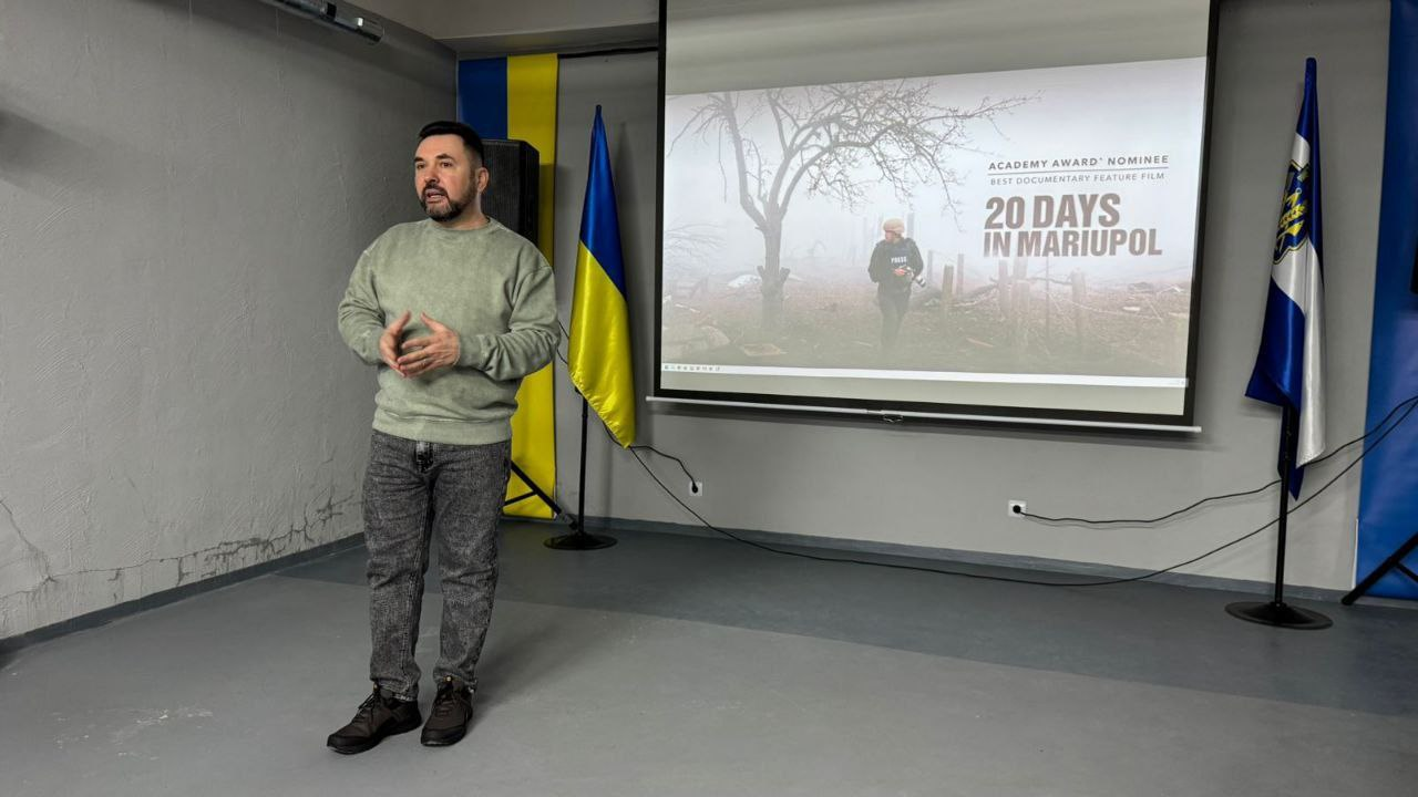 Чоловік виступає перед аудиторією на фоні великого екрана, на якому відображено постер документального фільму «20 днів у Маріуполі». Поруч із ним встановлено державний прапор України та офіційний стяг області.