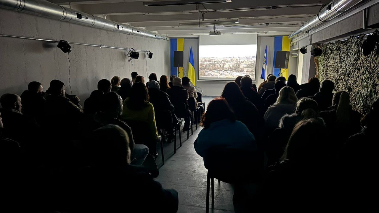 Глядачі в залі під час перегляду відео на великому екрані. Люди сидять рядами в напівтемному приміщенні, облаштованому в сучасному стилі зі світлими стінами та видимими елементами вентиляції. По обидва боки від екрана встановлені державні прапори України.