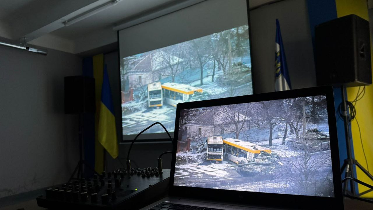 На передньому плані — відкритий ноутбук, що дублює зображення на великому екрані позаду. На екранах видно кадри з військовою технікою на засніженій вулиці міста. У приміщенні встановлені державні прапори України та аудіообладнання для проведення презентації.