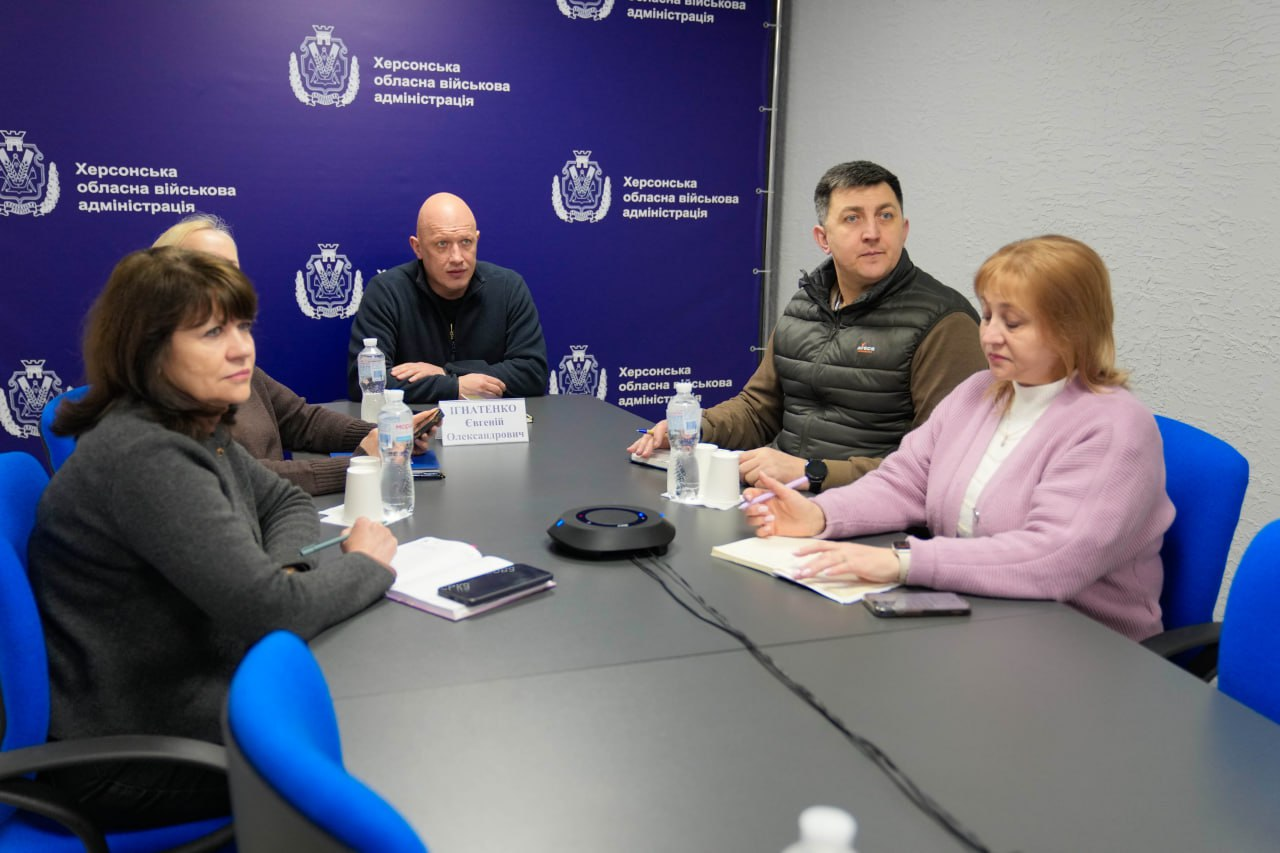 Робоча зустріч у кабінеті на фоні банера Херсонської обласної військової адміністрації. П'ятеро учасників сидять за столом, на якому розміщені блокноти, телефони, пляшки з водою та пристрій для гучного зв'язку. Присутні зосереджено слухають або ведуть записи.