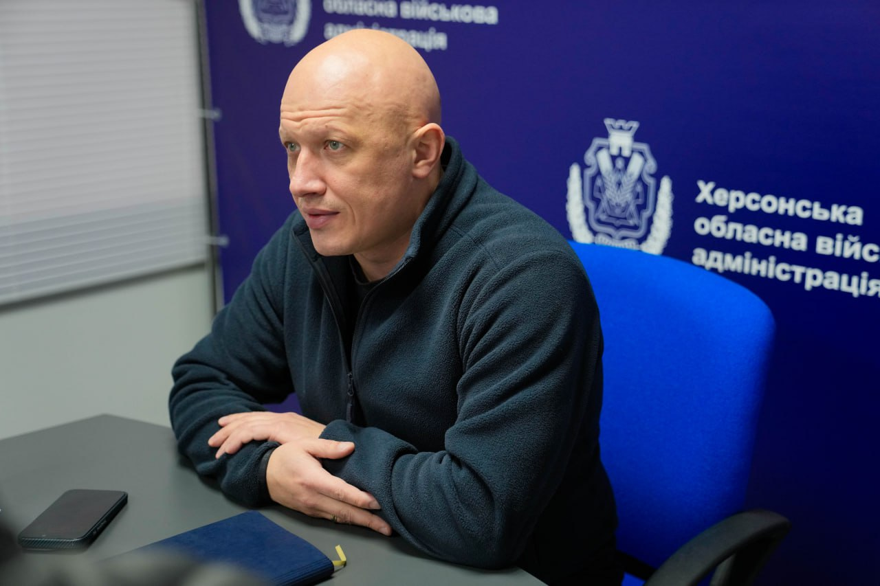 Чоловік у темній флісовій кофті сидить за робочим столом на фоні брендованого банера Херсонської ОВА. Перед ним на столі лежать закритий записник і смартфон.