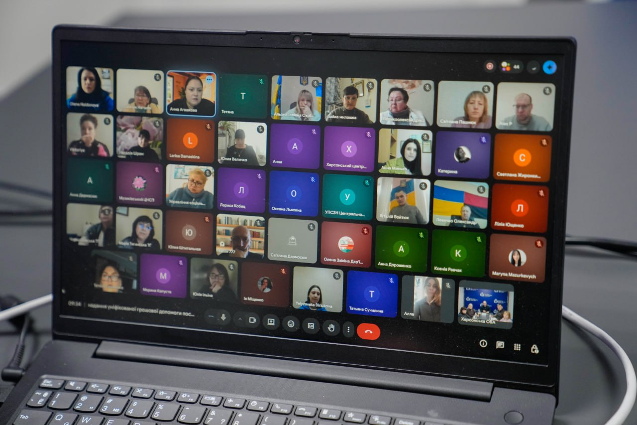 На екрані ноутбука відображено інтерфейс онлайн-наради з великою кількістю учасників у форматі відеоконференції. Видно вікна з відео та іконки з ініціалами користувачів; один із учасників транслює відео на фоні прапора України.