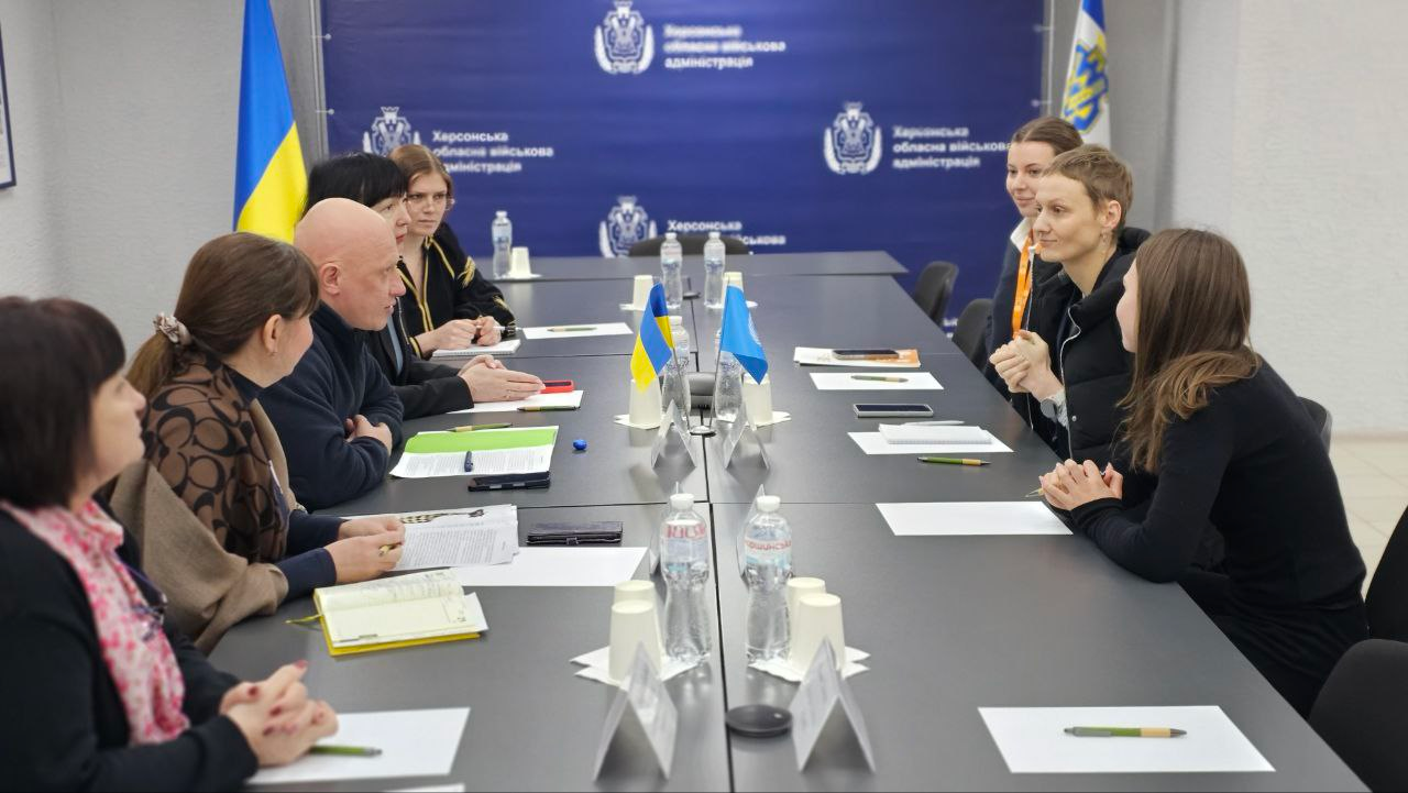 Робоча зустріч двох делегацій за довгим столом у кабінеті з символікою Херсонської ОВА. Учасники сидять один навпроти одного, ведуть діалог та роблять записи; на столі розміщені маленькі прапори України та ООН, документи й пляшки з водою.