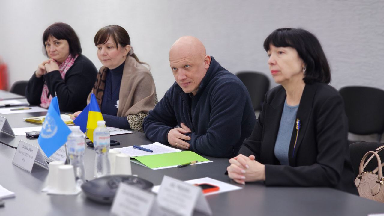 Чотири людини сидять за робочим столом під час офіційної зустрічі. На столі перед ними розміщені маленькі прапори України та ООН, документи в папках, пляшки з водою та пристрій для конференц-зв'язку. Учасники зосереджено слухають співрозмовників, які знаходяться навпроти.