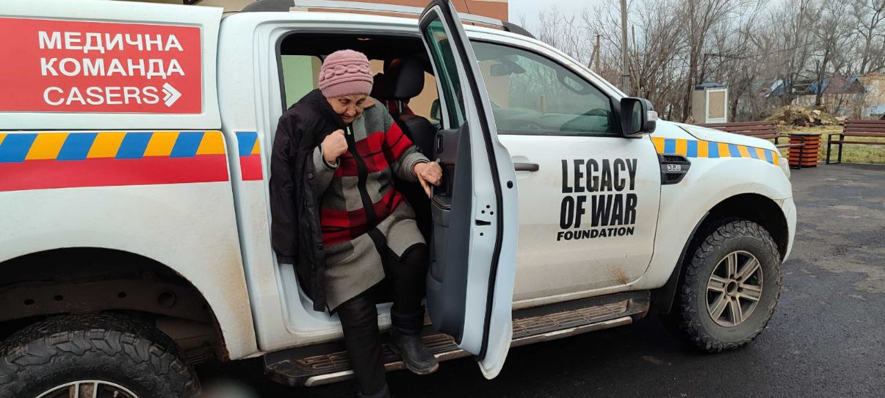 Жінка похилого віку виходить із білого позашляховика медичної команди «CASERS» та «Legacy of War Foundation». Автомобіль має спеціальне маркування та обладнаний для роботи в складних умовах.