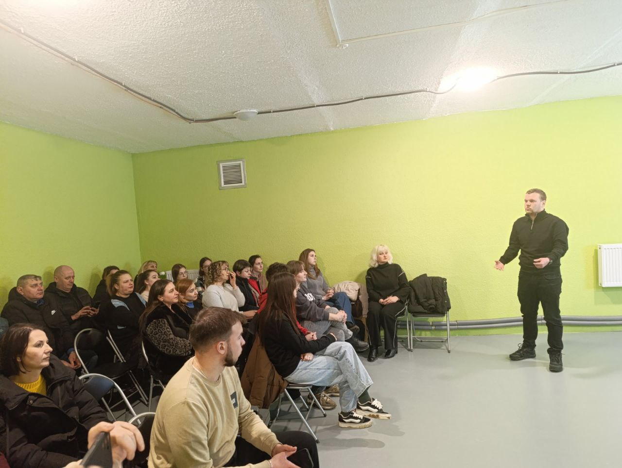 Чоловік у чорному одязі виступає перед аудиторією в залі з салатовими стінами. Слухачі, переважно молодь, уважно стежать за промовою спікера під час інформаційного заходу.