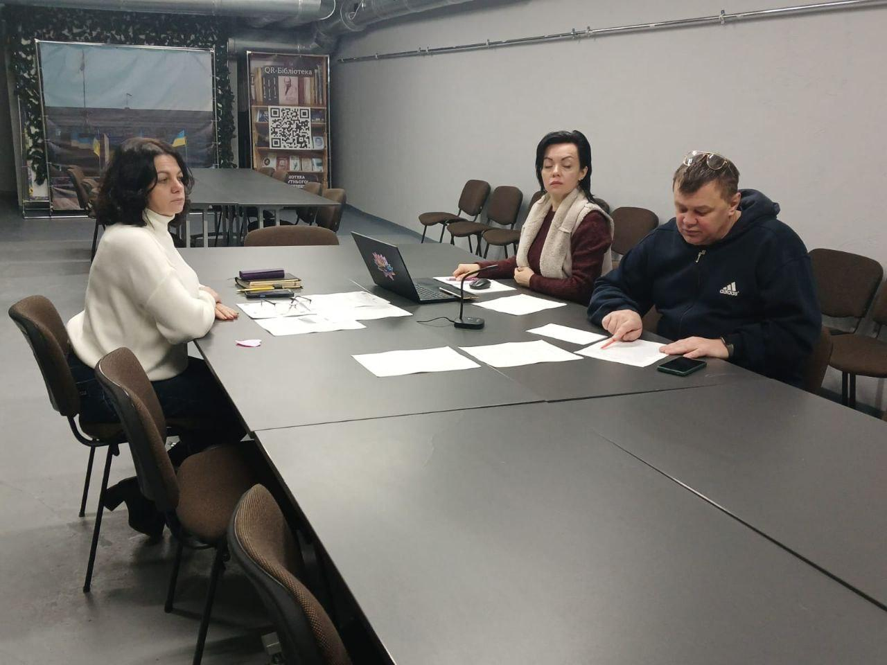 Робоча зустріч трьох осіб у конференц-залі. Учасники сидять за великим столом, працюють із документами та ноутбуком. На фоні видно банер із QR-кодом «Бібліотека».