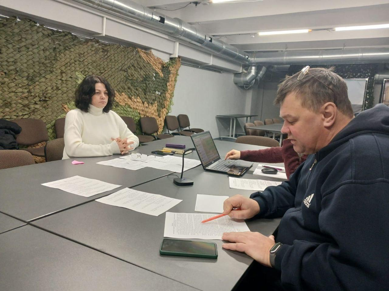 Троє людей проводять робочу зустріч у приміщенні. Вони сидять за столом, на якому лежать документи, ноутбук та мікрофон. На стіні позаду видно маскувальну сітку.