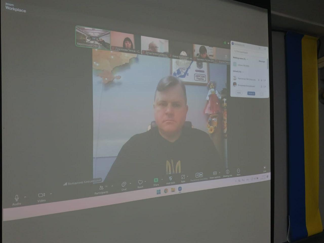 Фотографія екрана під час онлайн-конференції у Zoom. На екрані видно вікна з учасниками відеозустрічі, а праворуч від проекції розміщено державний прапор України.