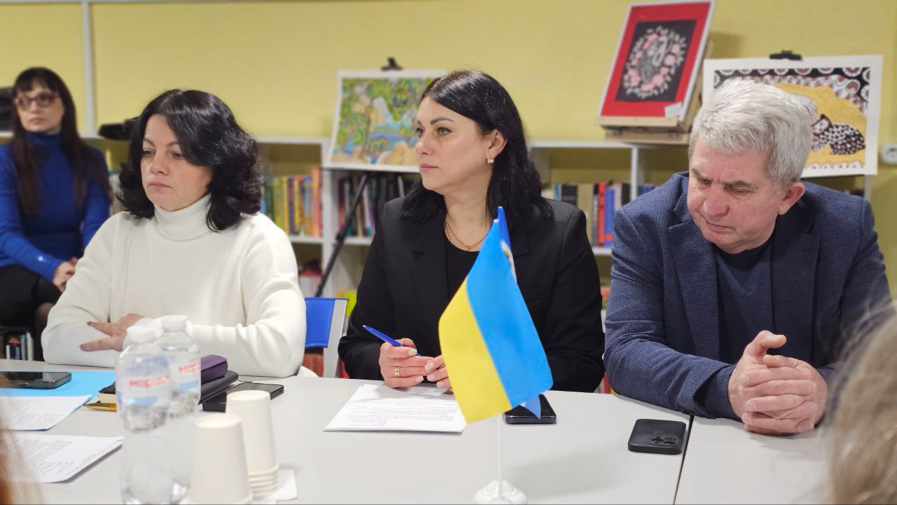 Троє учасників зустрічі за столом із державним прапорцем на передньому плані. На задньому плані видно ще одну особу, книжкові полиці та виставлені художні роботи.