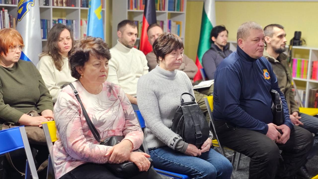 Група людей сидить на стільцях під час офіційного заходу. На задньому плані видно книжкові полиці та державні прапори України та інших країн.