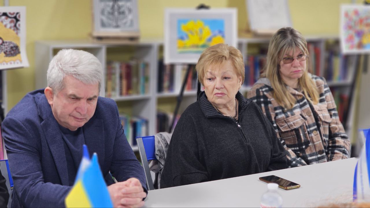 Троє людей сидять за столом під час заходу в бібліотеці чи культурному центрі. На фоні розташовані полиці з книгами та виставка дитячих малюнків, а на передньому плані видно частину державного прапора України.
