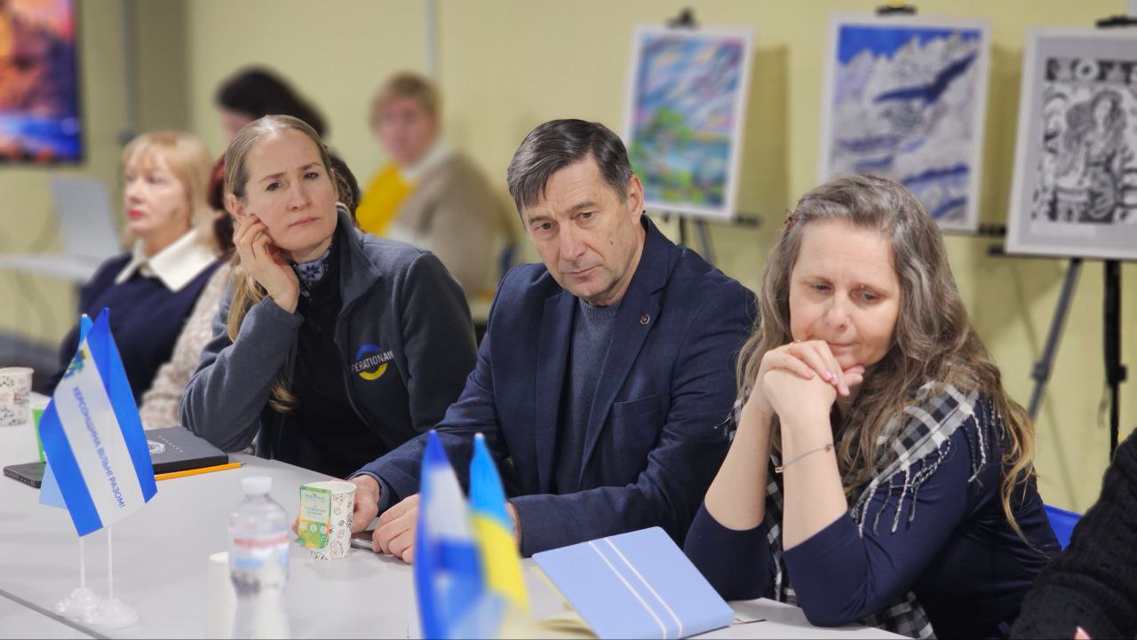 Четверо учасників зустрічі сидять за довгим столом, зосереджено слухаючи доповідача. Перед ними стоять настільні прапорці України та міжнародних організацій, а на задньому плані розміщена виставка художніх малюнків на мольбертах.