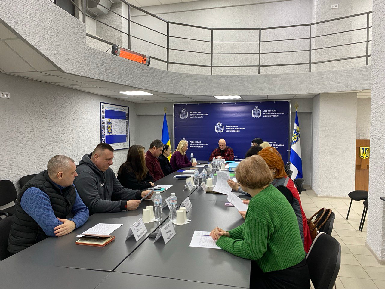 Загальний вид робочої зустрічі у просторій залі з балконом. Учасники засідання сидять навколо довгого столу на фоні синього банера з офіційною символікою, поруч із яким встановлені Державний Прапор України та прапор Херсонської області.