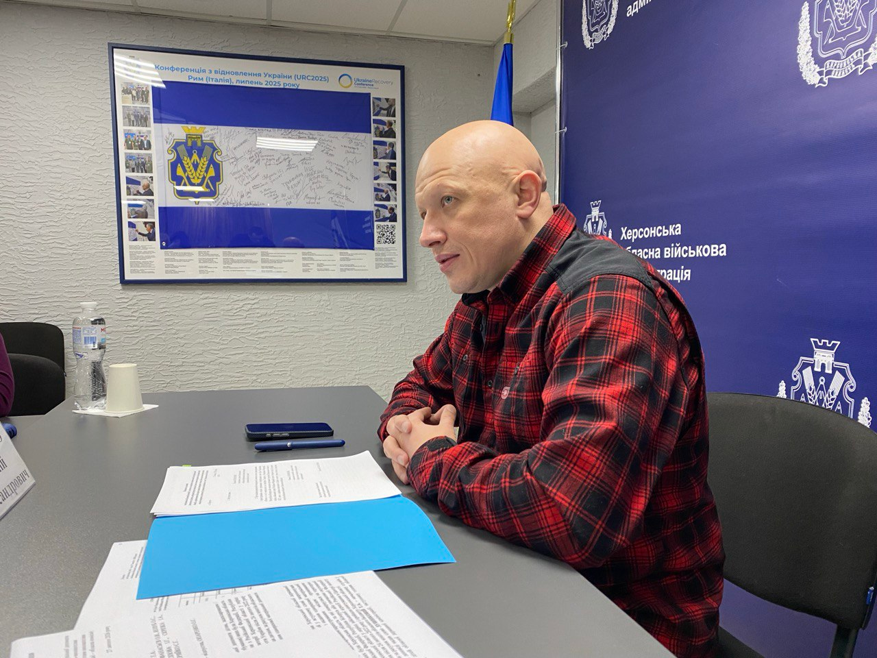 Чоловік у червоній клітчастій сорочці сидить за робочим столом під час офіційної зустрічі. На столі лежать документи, смартфон та ручка. На задньому плані — стіна з офіційною символікою Херсонської обласної військової адміністрації та плакат, присвячений Конференції з відновлення України.