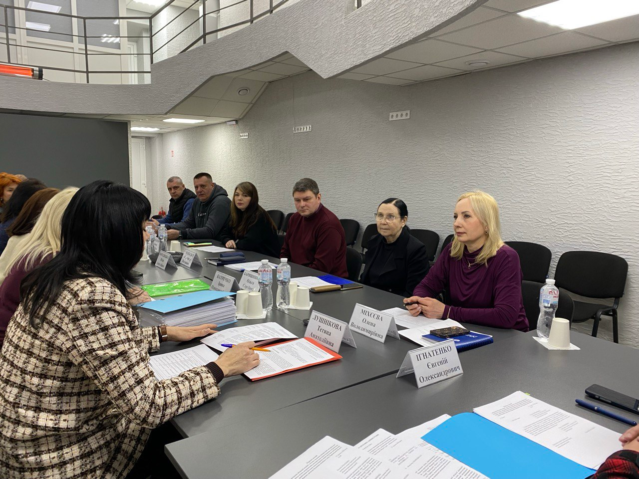 Група людей бере участь у робочій нараді за великим столом у сучасному офісному приміщенні. Перед учасниками розкладені документи, блокноти та таблички з іменами. Атмосфера зосереджена, присутні вивчають папери та обговорюють робочі питання.