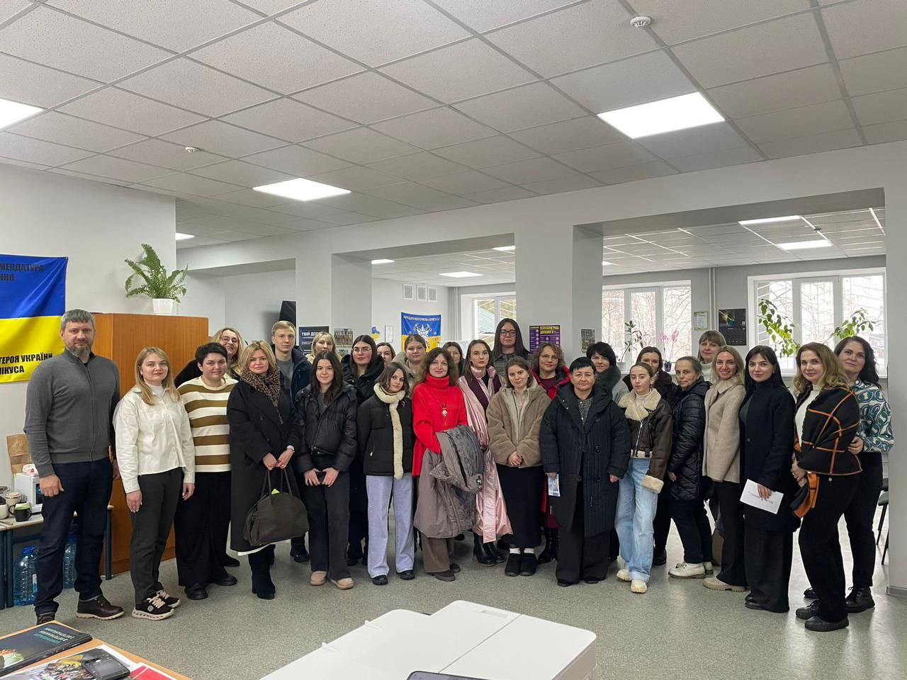 Велика група людей стоїть разом для спільного фото у просторому світлому залі. У приміщенні видно державну символіку України, інформаційні стенди та кімнатні рослини. Учасники одягнені у повсякденний та діловий одяг.