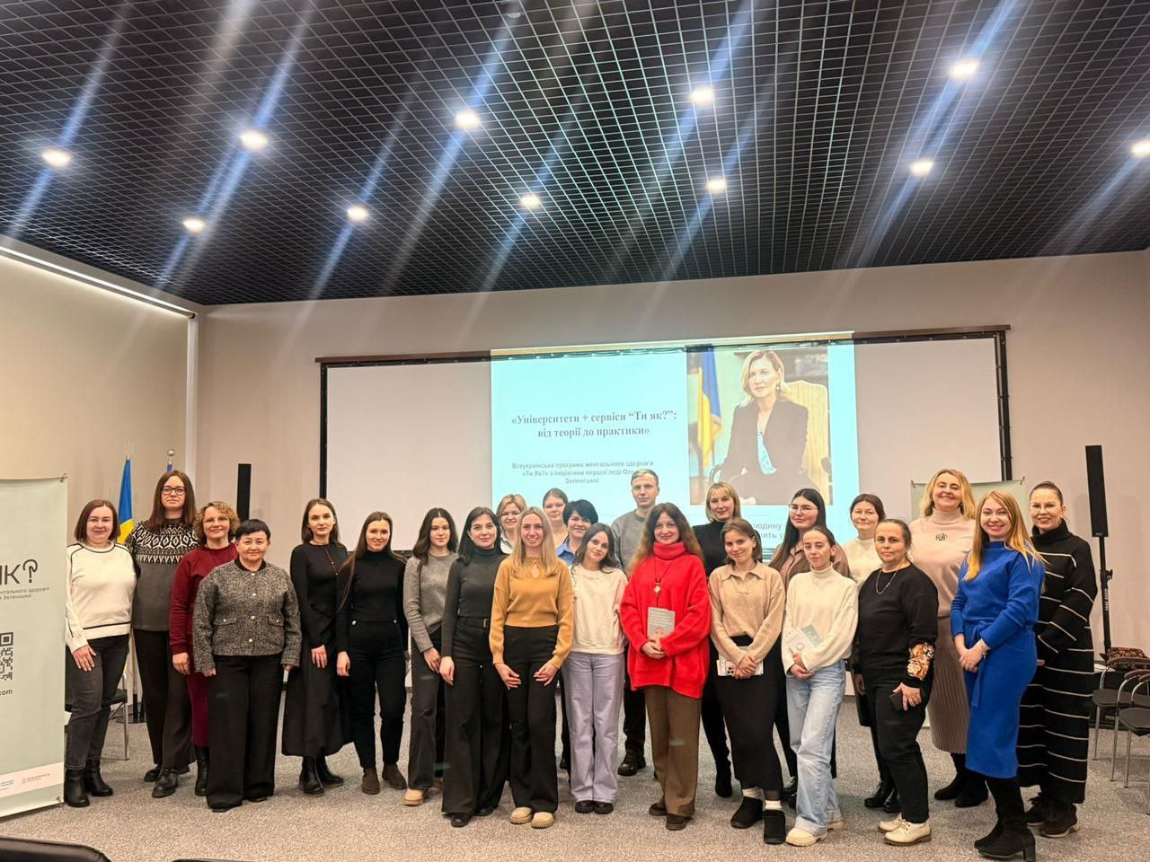 Група людей позує для фото на фоні великого екрана з презентацією про ментальне здоров'я. Зал має сучасне точкове освітлення та лаконічний інтер'єр. Учасники стоять у кілька рядів, деякі тримають у руках інформаційні буклети.