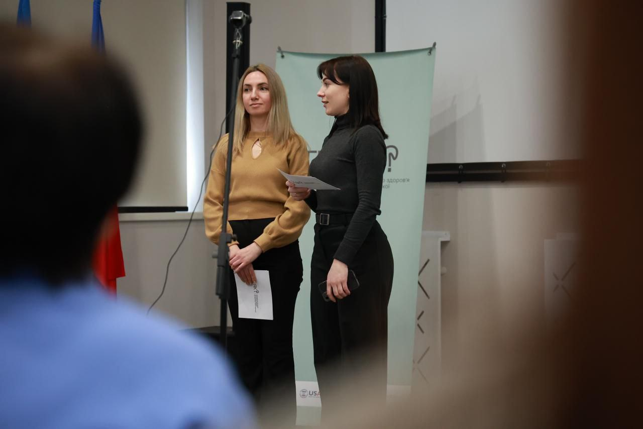 Дві жінки стоять перед аудиторією під час офіційного заходу. Одна з них тримає мікрофон, інша — інформаційні матеріали. На задньому плані видно частину банера з логотипом програми ментального здоров'я та прапор України.