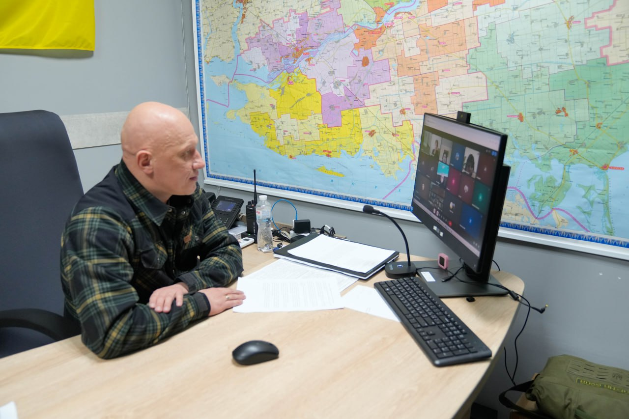 Чоловік у клітчастій сорочці сидить за робочим столом перед монітором комп'ютера, на якому триває відеоконференція з багатьма учасниками. На стіні за ним розміщена велика детальна карта області, а поруч — прапор України. На столі лежать робочі документи та мікрофон.