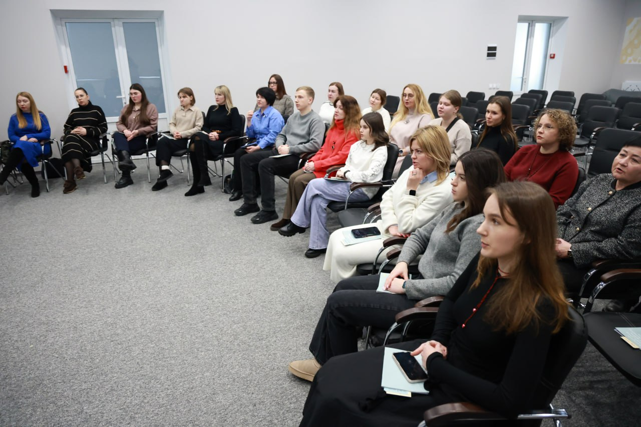 Група людей сидить на стільцях, розставлених півколом у світлій сучасній аудиторії. Учасники зосереджено дивляться в один бік, слухаючи спікера. Всі одягнені у повсякденний одяг, атмосфера заходу робоча та уважна.