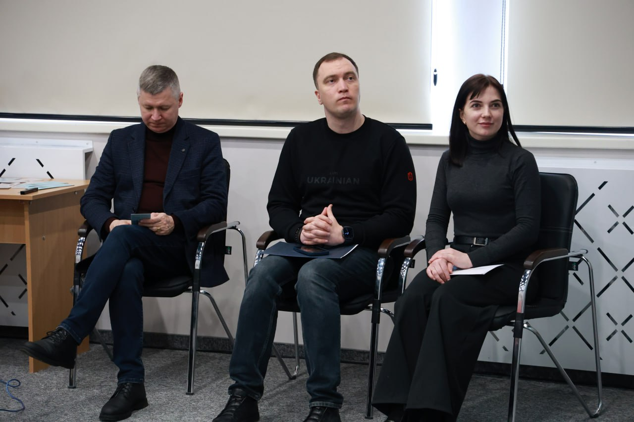 Троє учасників заходу — двоє чоловіків та жінка — сидять поруч на офісних стільцях під час робочої зустрічі. Вони одягнені в темний лаконічний одяг і зосереджено дивляться в бік спікера. На фоні видно частину світлої стіни та робочий стіл із документами.