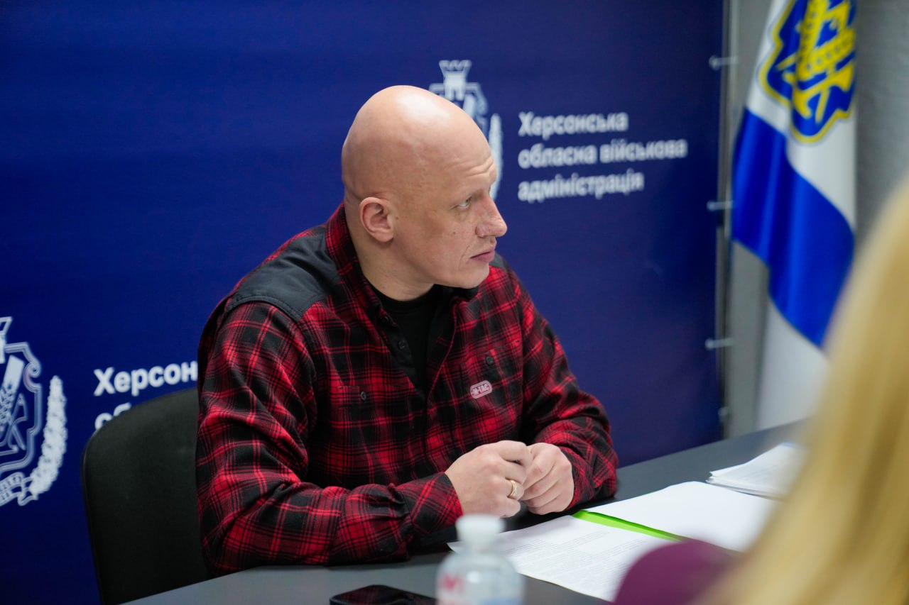 Крупний план чоловіка в червоній клітчастій сорочці, який зосереджено бере участь у робочій нараді. Він сидить за столом на фоні синього банера з офіційним логотипом Херсонської обласної військової адміністрації.