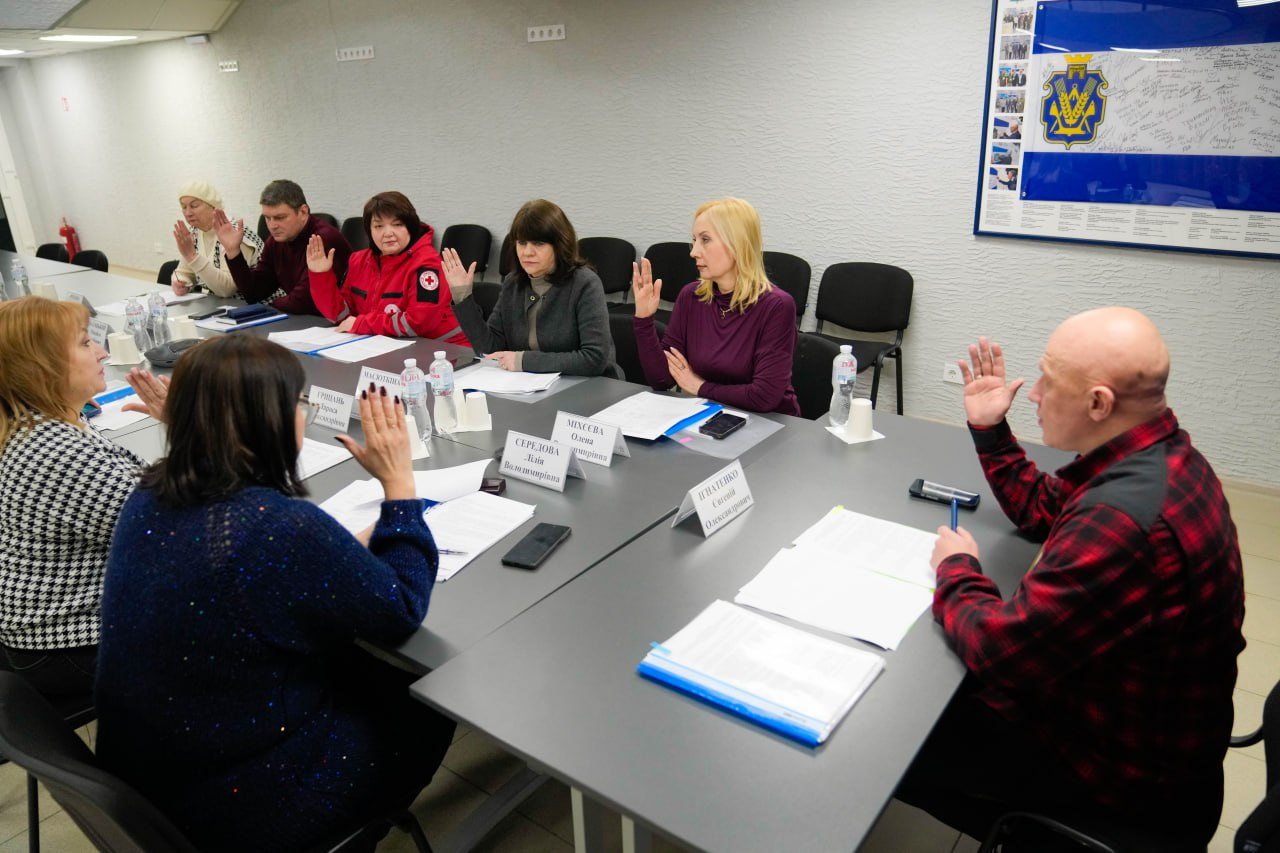Учасники робочої зустрічі в залі Херсонської ОВА одностайно голосують, піднявши руки. Вони сидять за великим столом із документами та табличками з прізвищами. На стіні праворуч розміщено стенд із державною символікою та фотографіями.