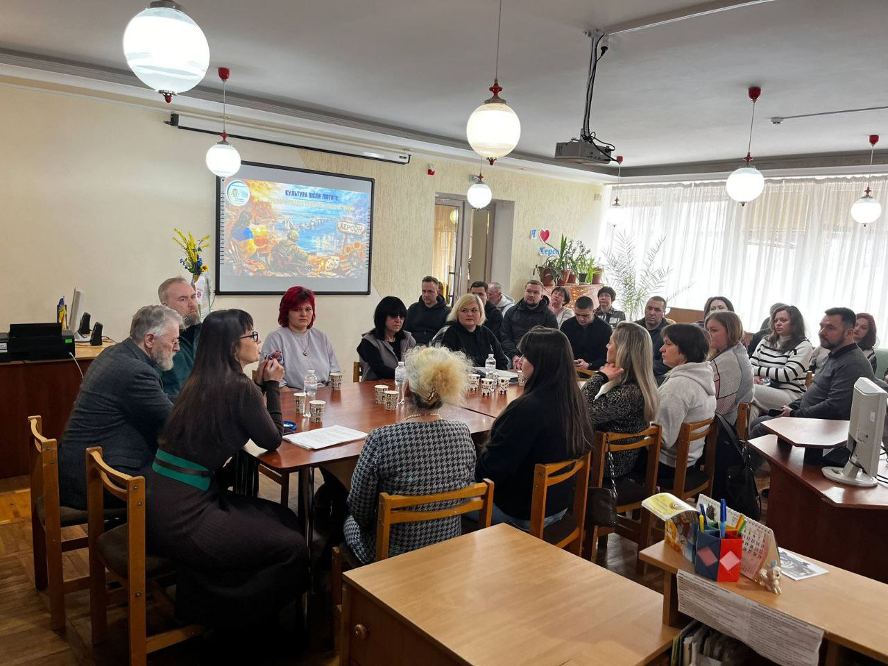 Група людей сидить за великим столом у світлій залі під час зустрічі чи презентації. У центрі уваги — екран із зображенням та написом «Культура після лютого», на якому видно патріотичну ілюстрацію з краєвидом Херсона. Учасники зосереджено слухають та обговорюють тему.