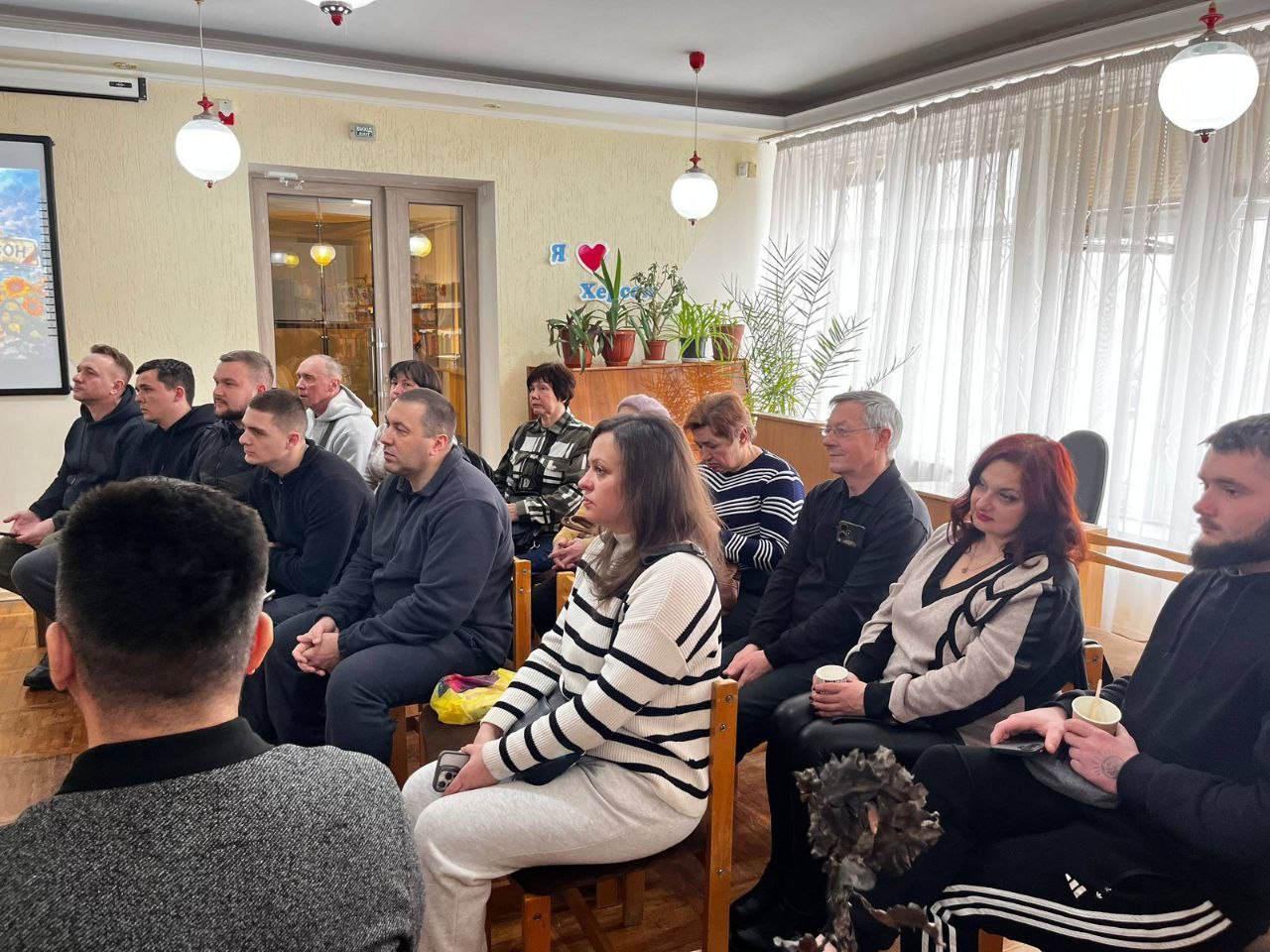 Група людей у приміщенні сидить рядами на стільцях, уважно слухаючи доповідача. Учасники зосереджені, дехто тримає паперові стаканчики з напоями. На задньому плані видно кімнатні рослини на підвіконні, світлі штори та частину екрана з презентацією. Атмосфера робоча та спокійна.