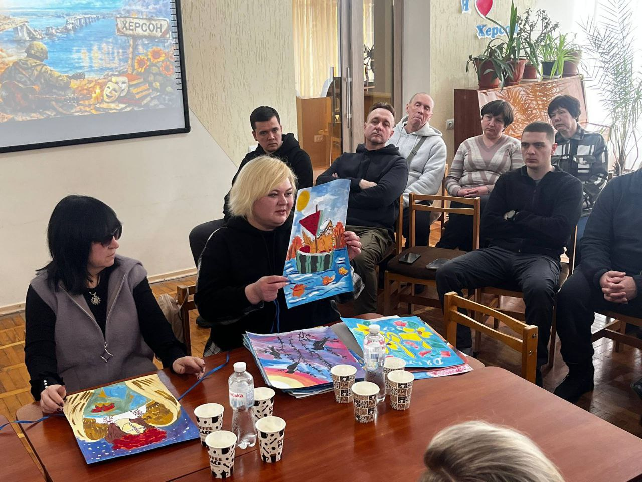 Жінка за столом демонструє аудиторії дитячий малюнок із зображенням кавуна-корабля на воді. На столі перед нею лежать інші кольорові малюнки та паперові стаканчики. Присутні в залі спостерігають за презентацією, на фоні видно екран із патріотичним плакатом про Херсон.