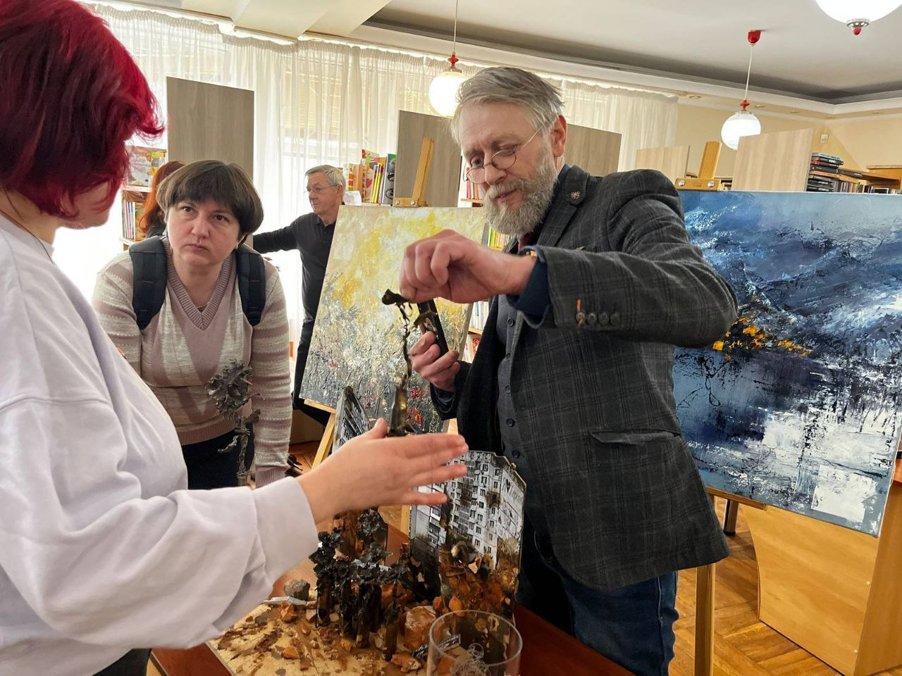 Чоловік у піджаку демонструє іншим учасникам елемент обвугленої художньої інсталяції, що нагадує зруйновану будівлю. Поруч стоять зацікавлені слухачі. На фоні в художній студії чи бібліотеці розташовані великі абстрактні картини на мольбертах та книжкові полиці.