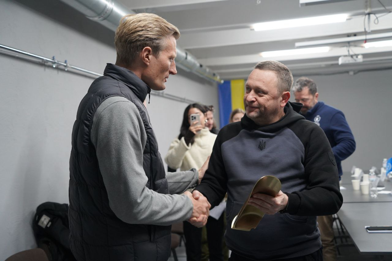 Двоє чоловіків тиснуть один одному руки під час зустрічі. Один із них тримає золотистий вигнутий предмет. На фоні присутні інші люди, одна із яких жінка, що робить фото на смартфон, також видніється прапор України.