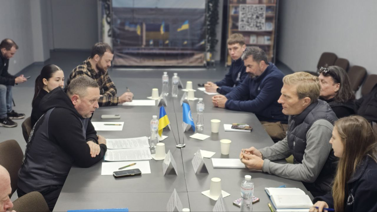 Група людей проводить робочу нараду за довгим столом у світлому приміщенні. У центрі столу встановлені маленькі прапорці України та Організації Об'єднаних Націй. Учасники обговорюють документи, роблять нотатки та використовують ноутбуки.