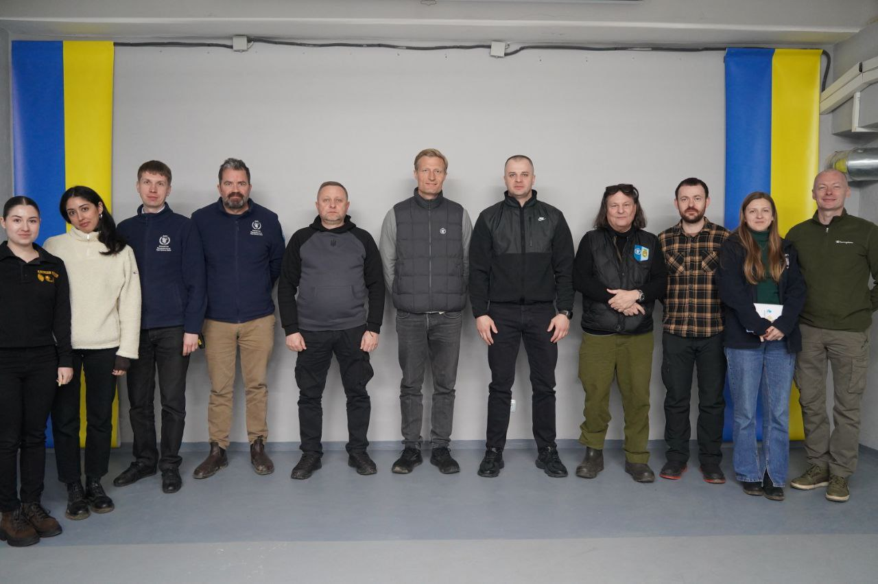 Групове фото одинадцяти учасників зустрічі, які стоять у ряд на фоні сірої стіни. По боках від групи розміщені два вертикальні прапори України. Учасники одягнені у зручний повсякденний одяг та куртки-жилетки.