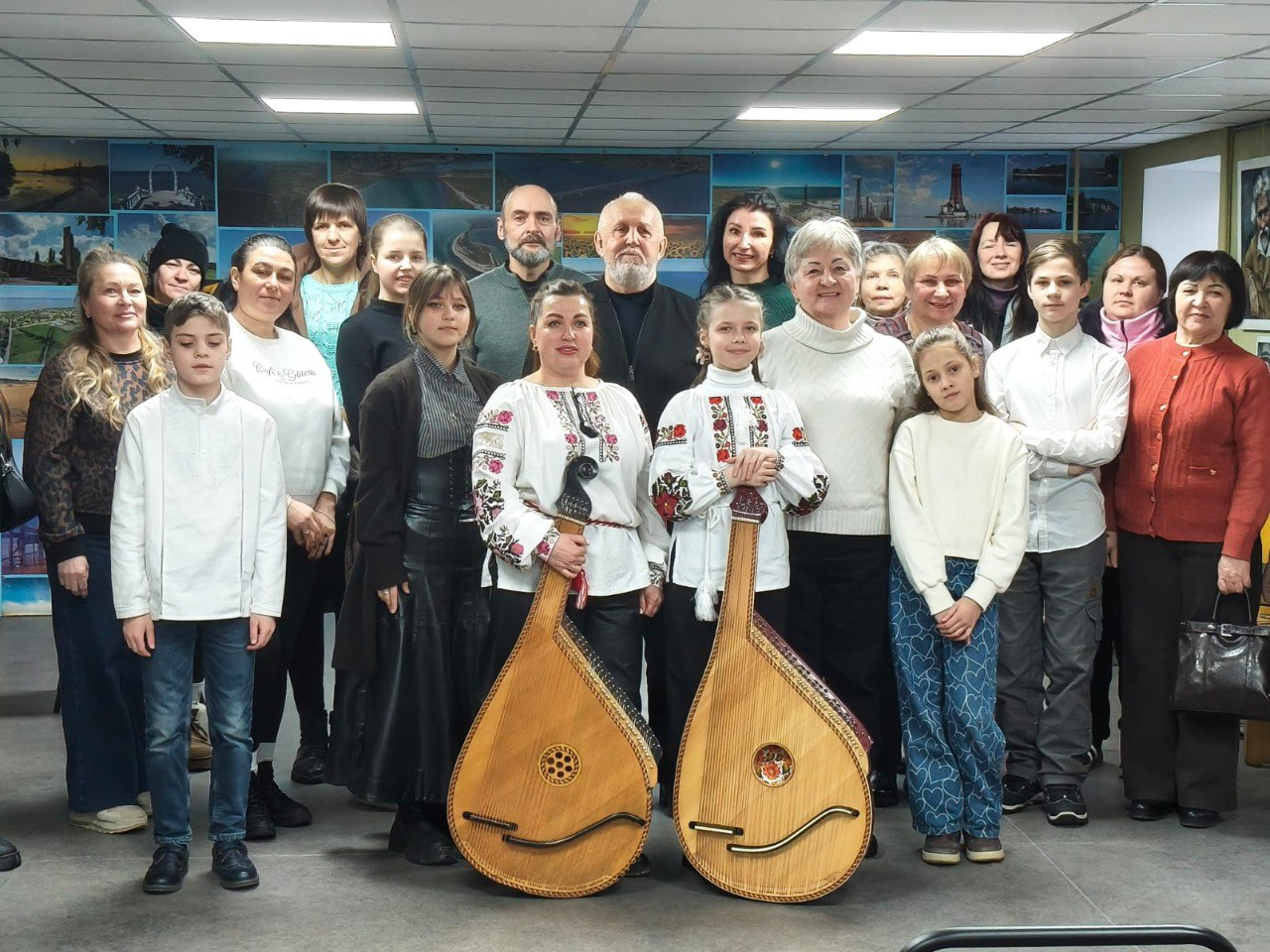 Групове фото учасників заходу в приміщенні. У центрі стоять двоє дівчат у вишиванках, кожна з яких тримає великий струнний музичний інструмент — бандуру. Навколо них розмістилися дорослі та діти у повсякденному одязі, а на задньому плані стіна прикрашена численними фотографіями краєвидів.