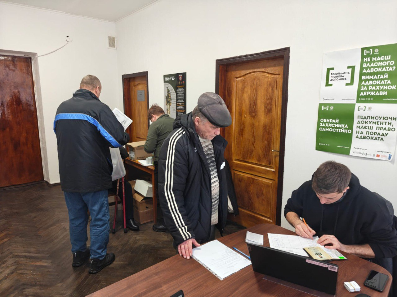 Чоловіки в коридорі та кабінеті установи займаються оформленням документів. Один чоловік стоїть біля столу та вивчає папери, інший заповнює бланк, сидячи за ноутбуком. На стіні розміщено інформаційні плакати про безоплатну правову допомогу.