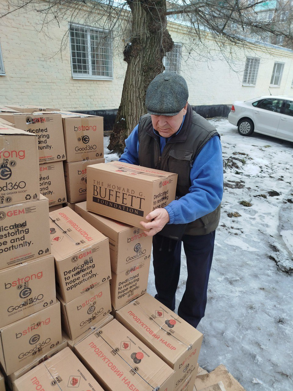 Літній чоловік у кепці та жилетці розвантажує коробки з гуманітарною допомогою надворі. Він тримає в руках картонний ящик із написом «The Howard G. Buffett Foundation», а поруч складено стоси аналогічних пакунків із позначками «Продуктовий набір».