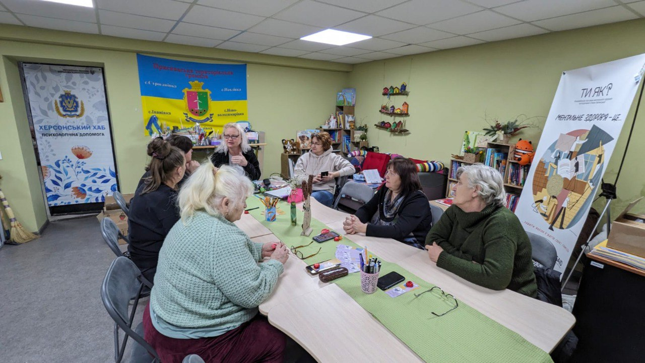Група жінок сидить за довгим столом у світлому кабінеті під час групового заняття або бесіди. У приміщенні розміщено стенд програми ментального здоров'я «Ти як?», прапор Присиваської територіальної громади та банер «Херсонський хаб: психологічна допомога». На столі лежать канцелярські приладдя та окуляри.