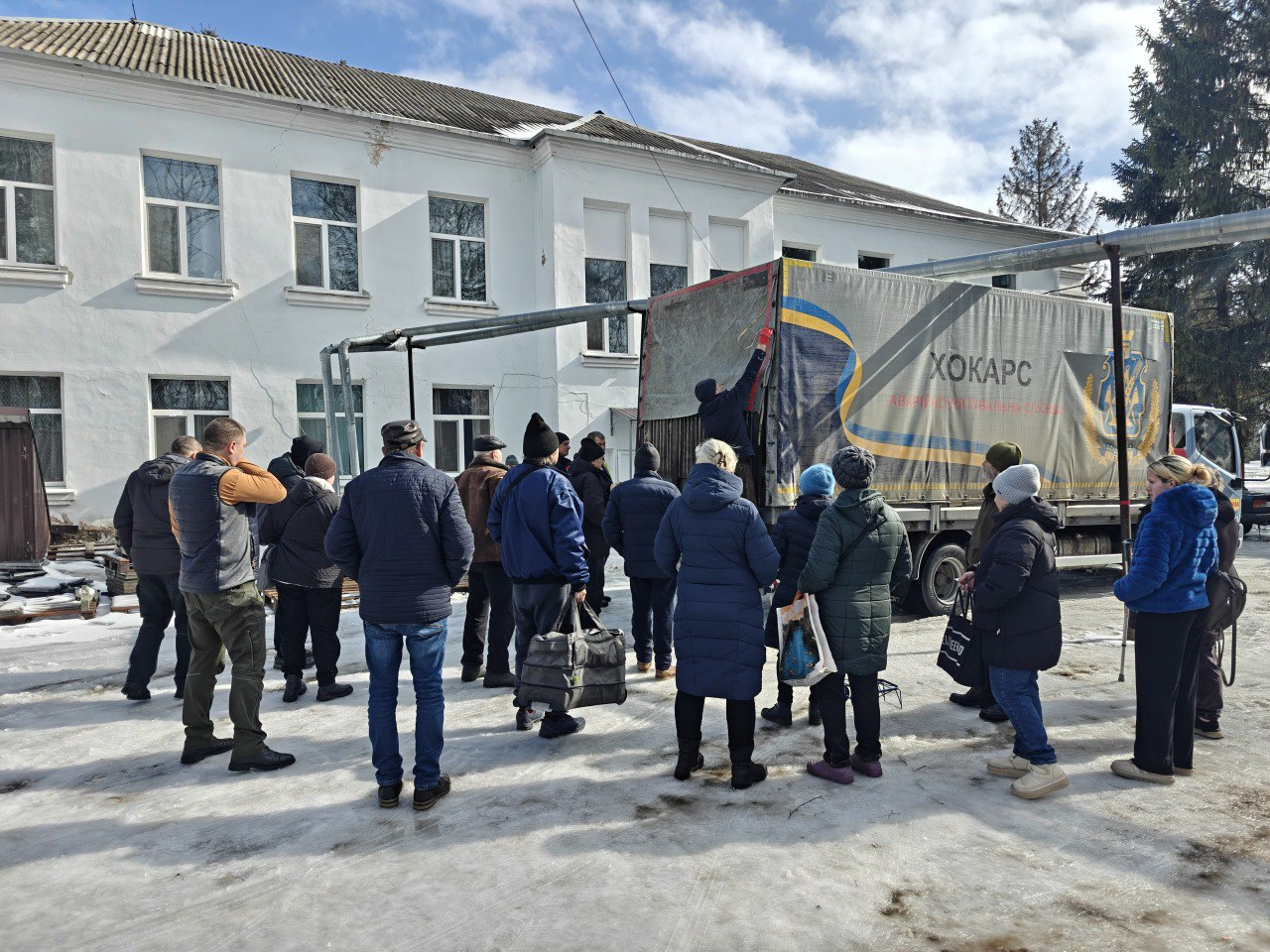На засніженому подвір'ї біля двоповерхової будівлі група людей чекає на розвантаження великої вантажівки. На тентованому кузові авто розміщено напис «ХОКАРС Аварійно-рятувальна служба» та емблему. Люди одягнені в теплий зимовий одяг і стоять спиною до камери.