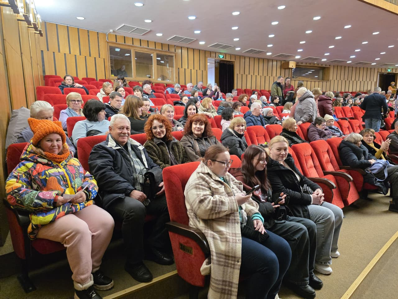 Глядацька зала з дерев'яним оздобленням стін, заповнена людьми різного віку. Учасники заходу сидять у рядах м'яких червоних крісел, деякі з них дивляться в камеру та посміхаються. Люди одягнені в різнобарвний повсякденний та верхній одяг.