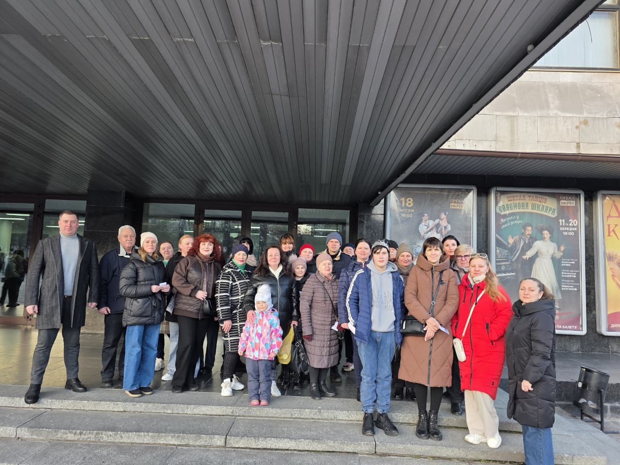 Велика група людей різного віку стоїть перед входом до будівлі театру чи будинку культури під широким козирком. Учасники заходу одягнені в зимовий та демісезонний верхній одяг. Праворуч на стіні розміщені афіші майбутніх вистав.