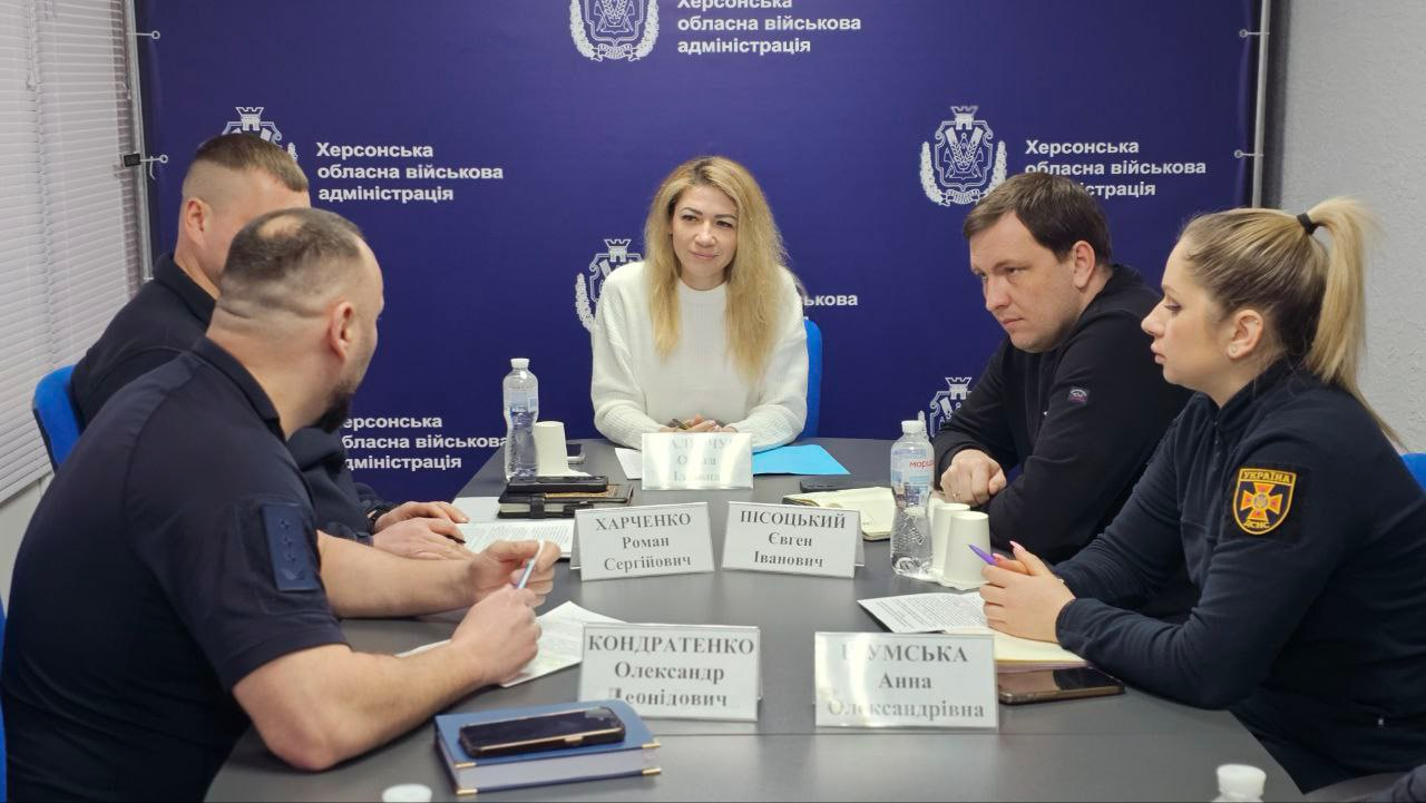 Робоча зустріч п'яти осіб у кабінеті за столом з іменними табличками. Серед присутніх — чоловіки та жінки в офіційному одязі та однострої рятувальної служби, які обговорюють робочі питання на фоні синього інформаційного стенда.